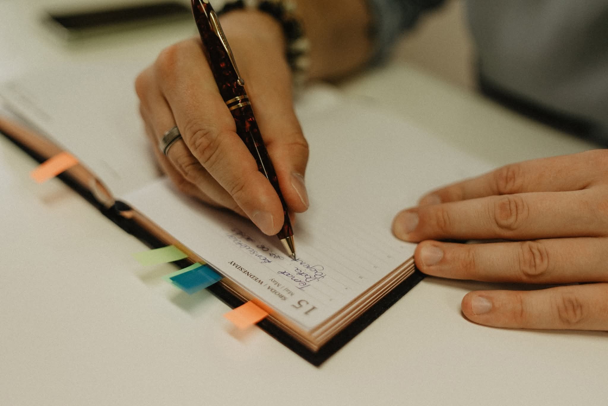 Osoba pisze w kalendarzu piórem z marmurkową obudową. Widoczne kolorowe zakładki, dzień tygodnia 'ŚRODA' i data '15 Maj'. Dłoń z pierścionkiem na palcu.