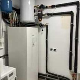 HELIOLINE - Biała pompa ciepła w kotłowni, z widocznymi rurami instalacji, zbiornikiem buforowym i pralką w tle. Ujęcie z niskiego kąta.