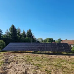 Długa instalacja paneli słonecznych na metalowej konstrukcji, umieszczona na lekko piaszczystym terenie z rzadką trawą, w tle drzewa i budynek z czerwoną dachówką pod błękitnym niebem.