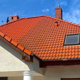 HELIOLINE - Czerwony dach z dachówki ceramicznej, widok z dołu na tle błękitnego nieba. Widoczny komin i okno dachowe. Elewacja budynku w jasnym kolorze.