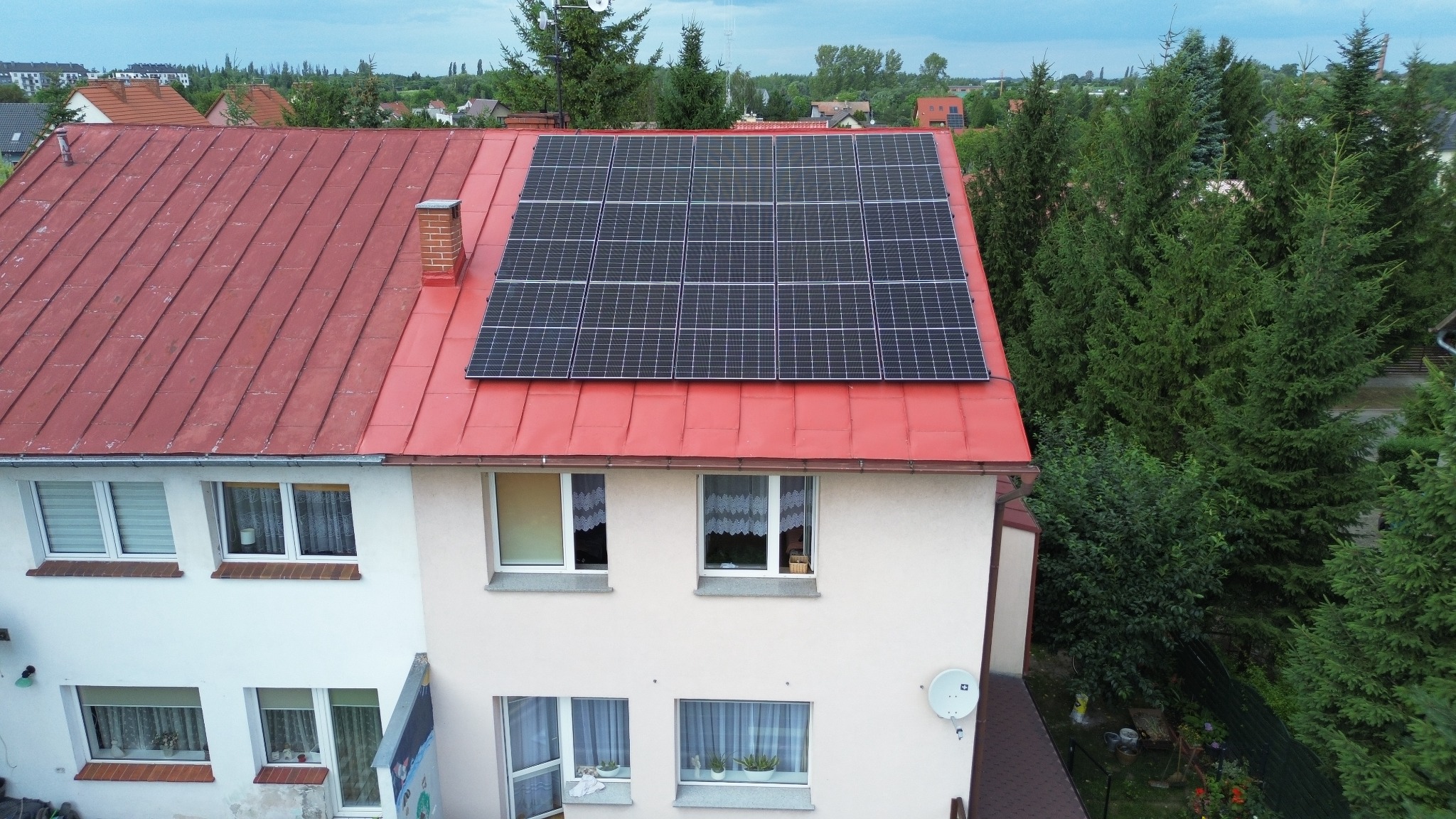 Panel słoneczny na czerwonym dachu dwukondygnacyjnego domu jednorodzinnego, widok z góry, otoczenie zieleni.