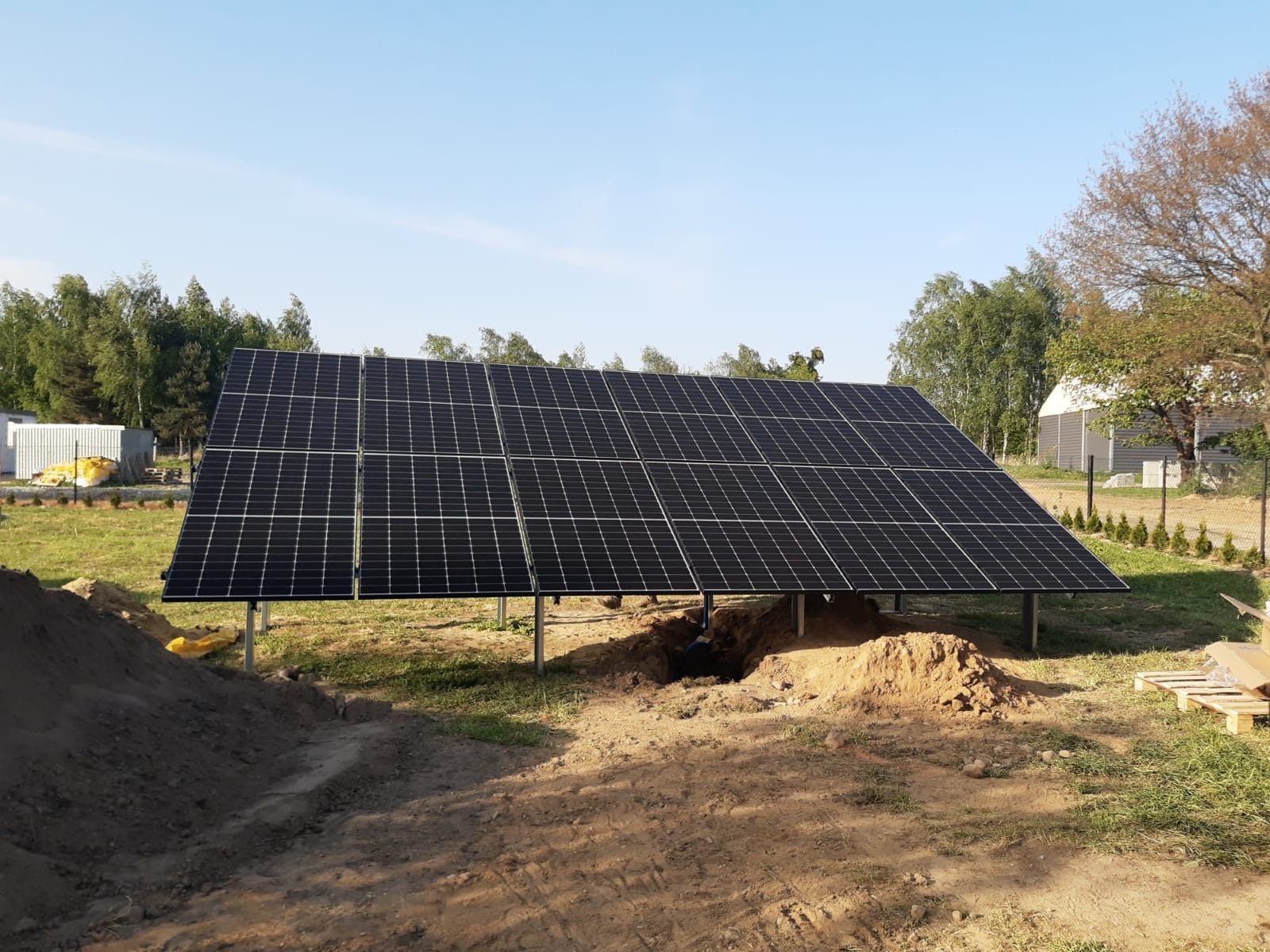 Instalacja paneli słonecznych na metalowych podporach, widoczne wykopy w ziemi i pryzma piasku, w tle budynki gospodarcze i drzewa.