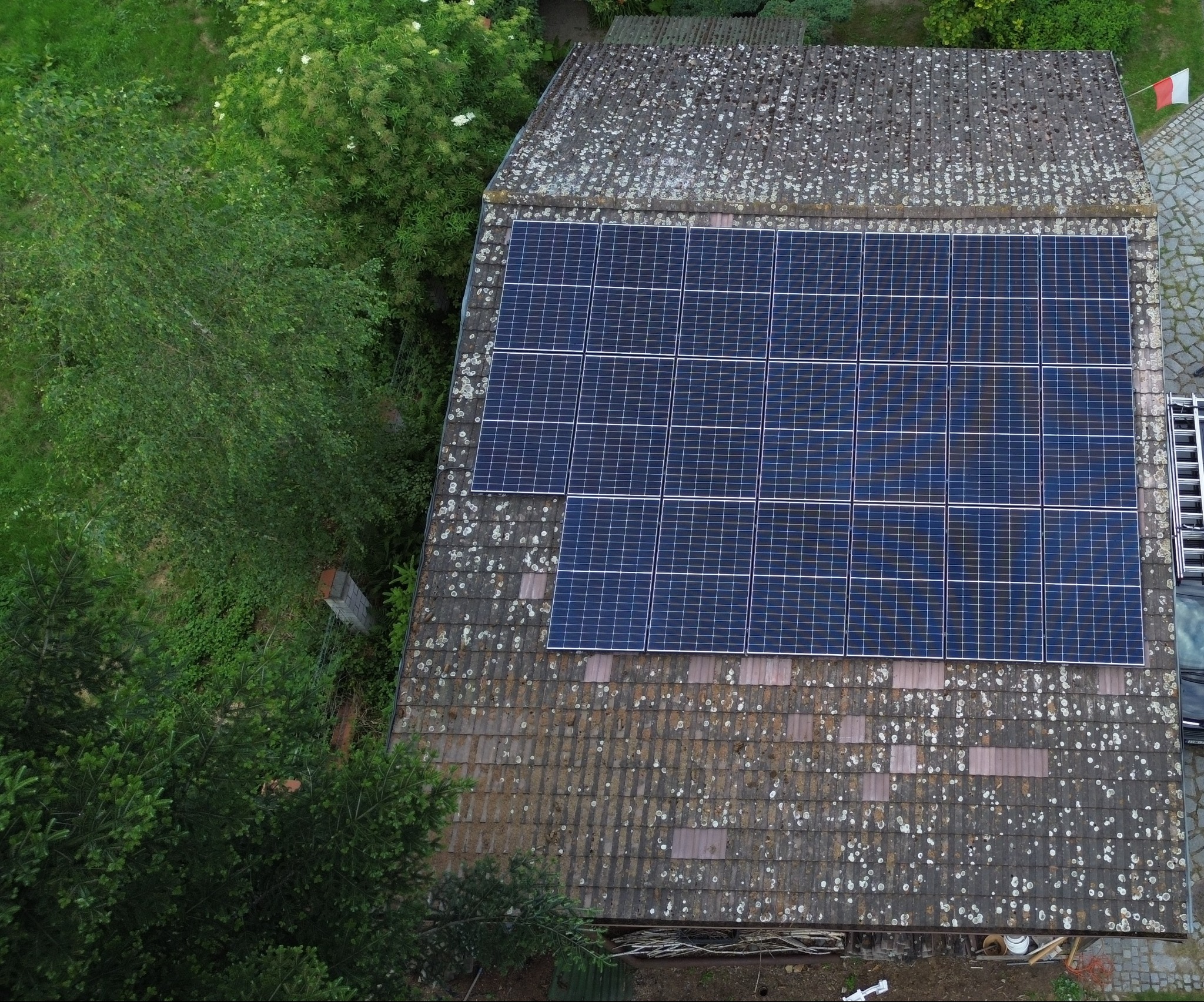 Widok z góry na instalację paneli słonecznych na zarośniętym mchem dachu domu we Wrocławiu, z widoczną drabiną i fragmentem samochodu obok. Na dachu powiewa flaga Polski.