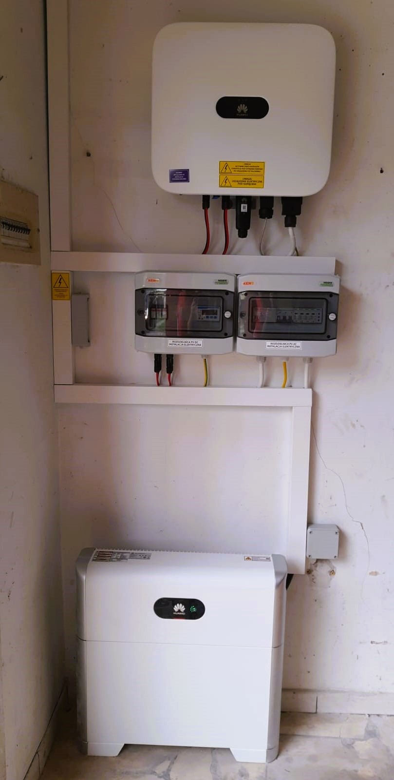 Biały magazyn energii Huawei obok białej ściany z zamontowanym inwerterem i rozdzielnicami elektrycznymi.