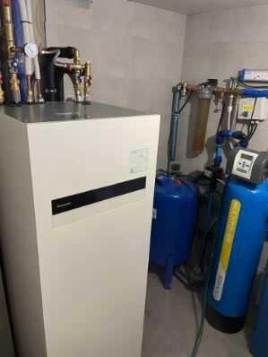 Biała pompa ciepła w kotłowni, widoczne niebieskie zbiorniki i rury instalacji, szare płytki na ścianie.