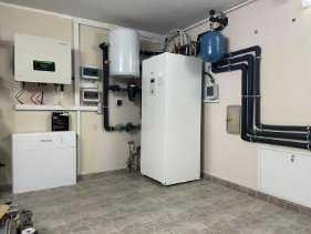 Biała pompa ciepła powietrze-woda w kotłowni z szarymi płytkami podłogowymi, widoczne rury instalacyjne, zbiornik wyrównawczy i inne urządzenia grzewcze.