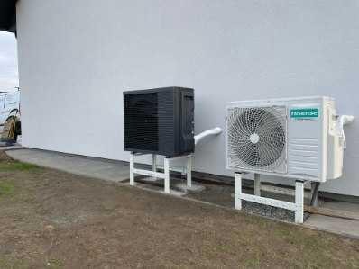 Dwie jednostki zewnętrzne pomp ciepła, czarna i biała, zamontowane na białych wspornikach przy białej ścianie budynku, widoczny fragment rury odprowadzającej skropliny z białej jednostki.