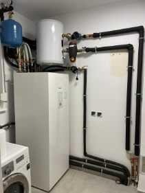 Biała pompa ciepła w kotłowni, z widocznymi rurami instalacji, zbiornikiem buforowym i pralką w tle. Ujęcie z niskiego kąta.