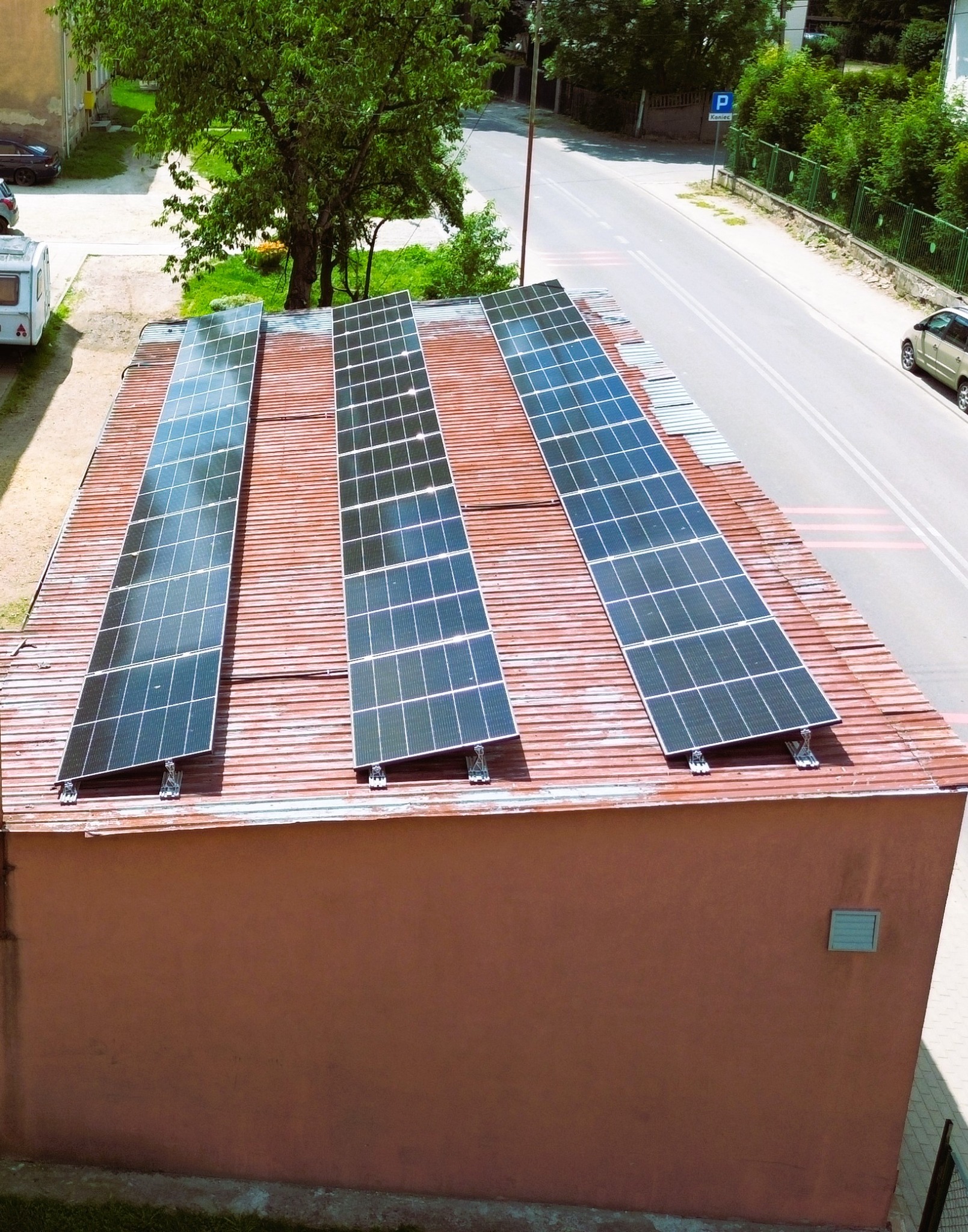Widok z góry na trzy rzędy paneli słonecznych zamontowanych na czerwonym, skośnym dachu budynku, z ulicą i zielenią w tle.
