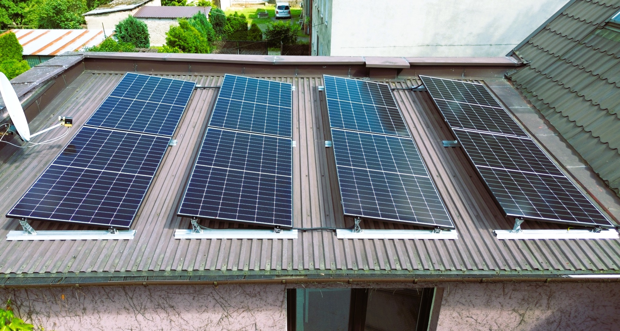 Cztery panele słoneczne zamontowane na metalowym dachu budynku z widoczną anteną satelitarną i fragmentem elewacji pokrytej bluszczem.