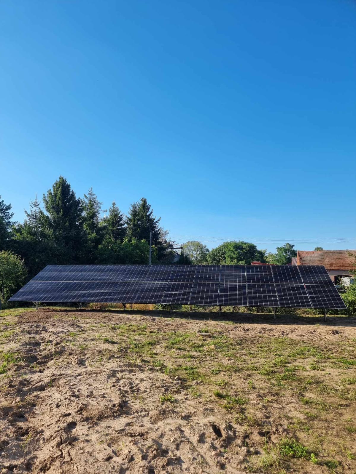 Długa instalacja paneli słonecznych na metalowej konstrukcji, umieszczona na lekko piaszczystym terenie z rzadką trawą, w tle drzewa i budynek z czerwoną dachówką pod błękitnym niebem.