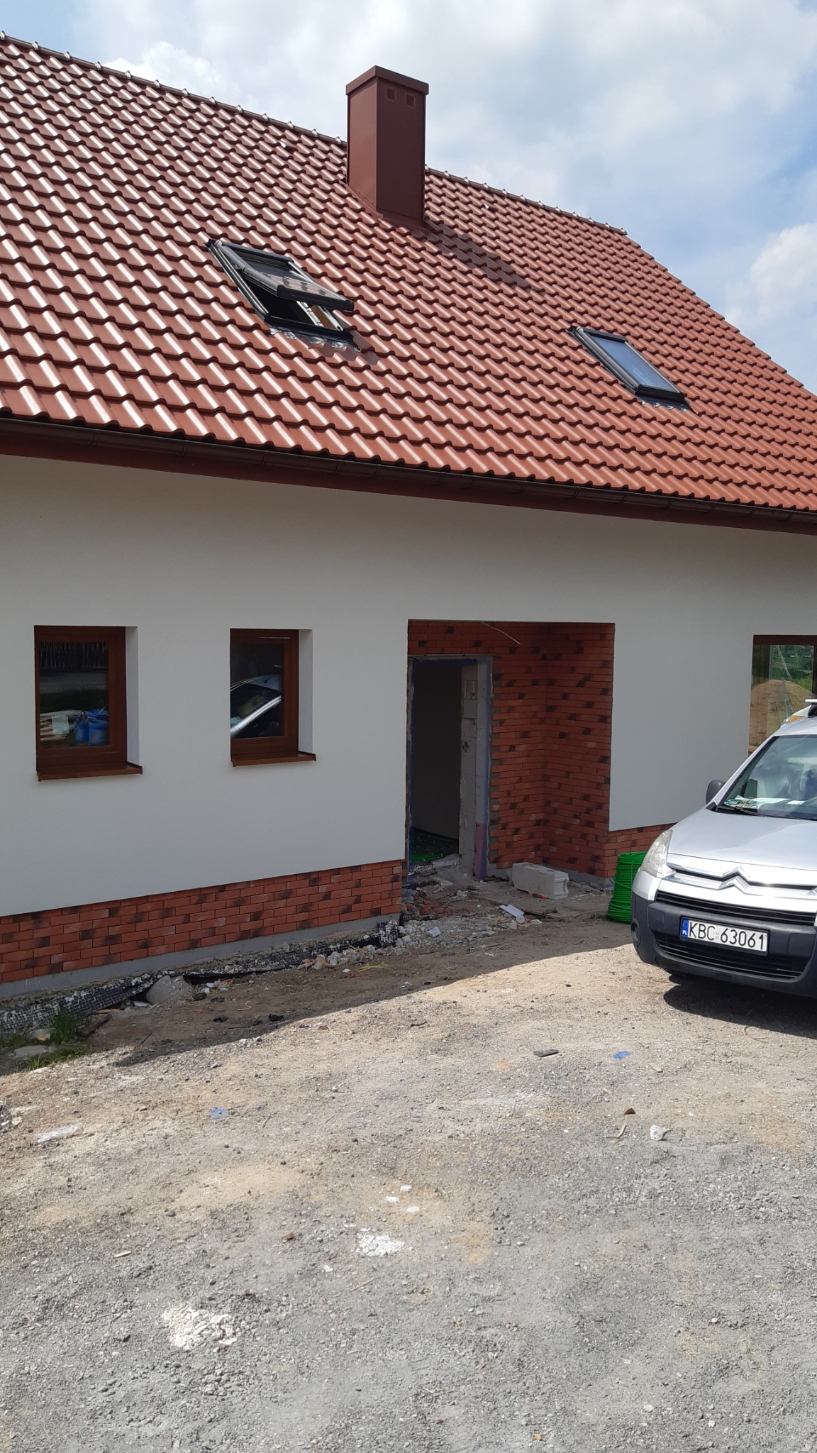 Dom jednorodzinny w trakcie wykańczania elewacji z cegły klinkierowej, widoczne okna z brązowymi ramami, dach z czerwonej dachówki z oknami połaciowymi i kominem, zaparkowany samochód dostawczy...
