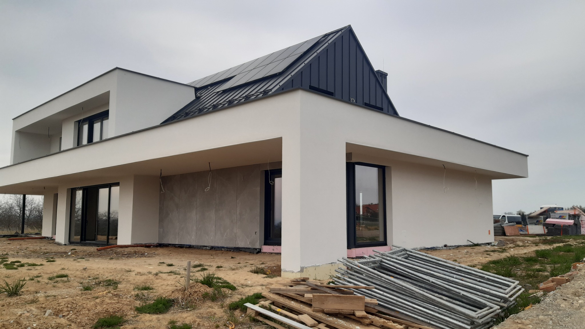 Nowoczesny dom jednorodzinny z płaskim dachem i panelami słonecznymi, w trakcie wykończenia elewacji, widoczne rusztowania i materiały budowlane na niezagospodarowanym terenie.