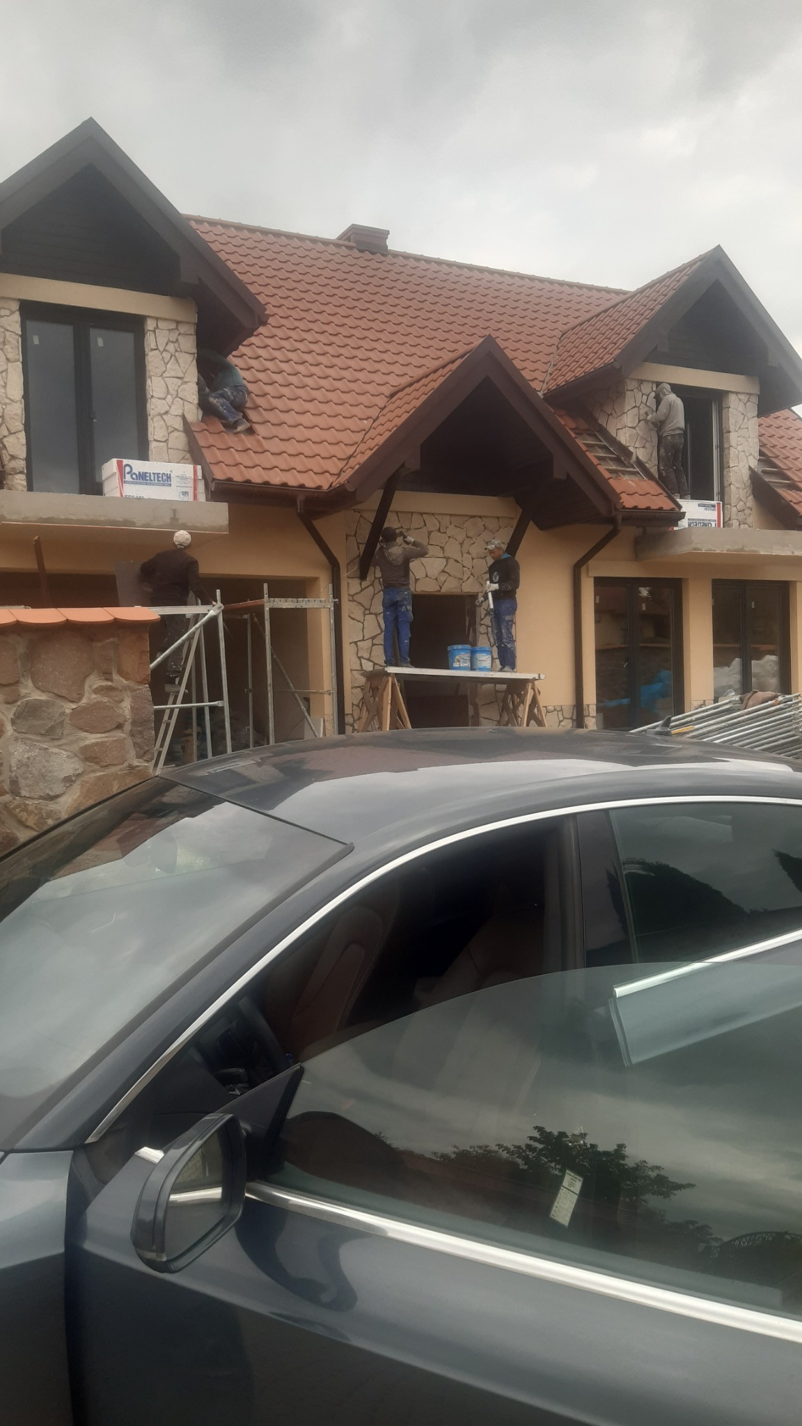 Prace wykończeniowe elewacji domu jednorodzinnego z kamienną okładziną, widoczni pracownicy na rusztowaniach i podestach podczas nakładania tynku, z przodu widoczny fragment samochodu.