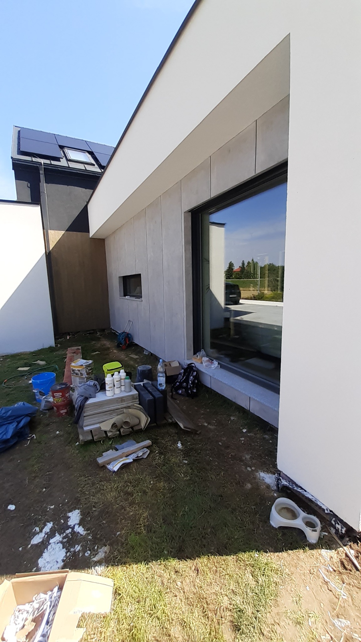 Fragment ocieplonej elewacji nowoczesnego domu z dużym oknem, widoczne materiały budowlane i narzędzia na trawie, w tle panele słoneczne na dachu.
