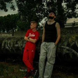 Dwóch młodych mężczyzn pozuje na tle betonowej rury z graffiti, jeden w czerwonym dresie, drugi w szarych spodniach cargo i czarnym podkoszulku, obaj z zegarkami na rękach.