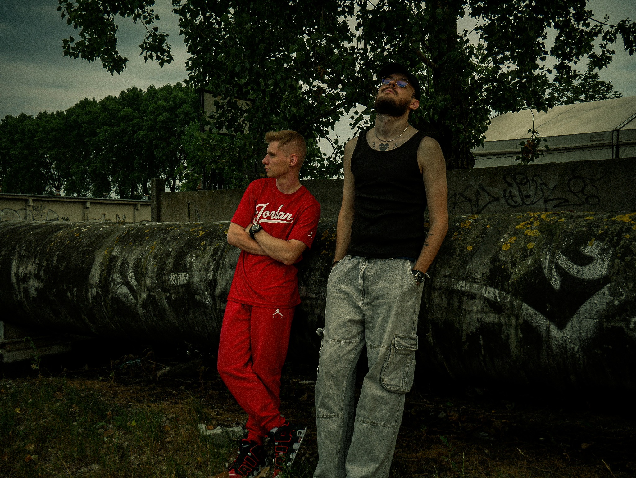 Dwóch młodych mężczyzn pozuje na tle betonowej rury z graffiti, jeden w czerwonym dresie, drugi w szarych spodniach cargo i czarnym podkoszulku, obaj z zegarkami na rękach.