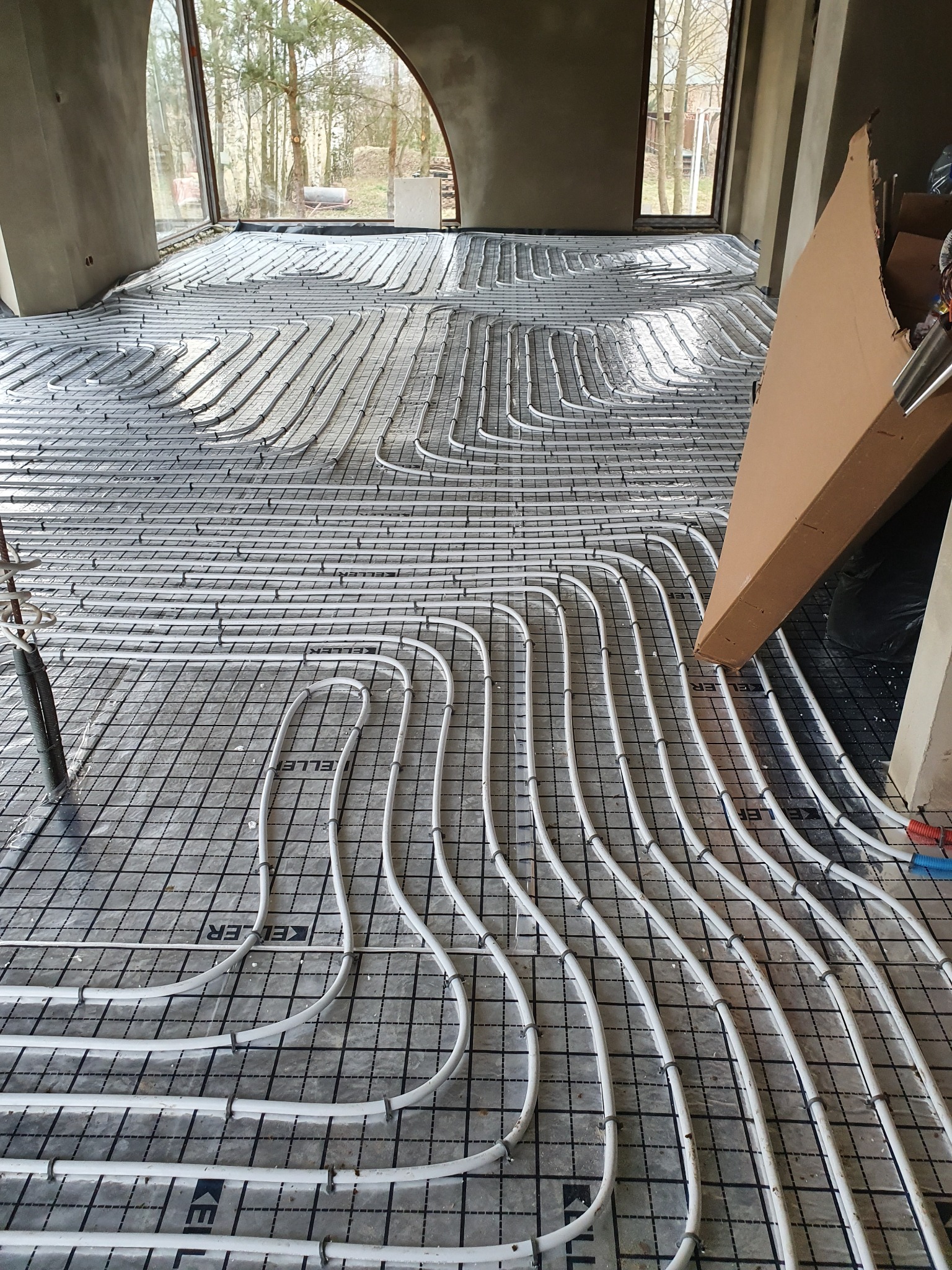 Białe rurki ogrzewania podłogowego ułożone w pętle na czarnej macie z siatką w surowym wnętrzu z dużym oknem i widokiem na drzewa.