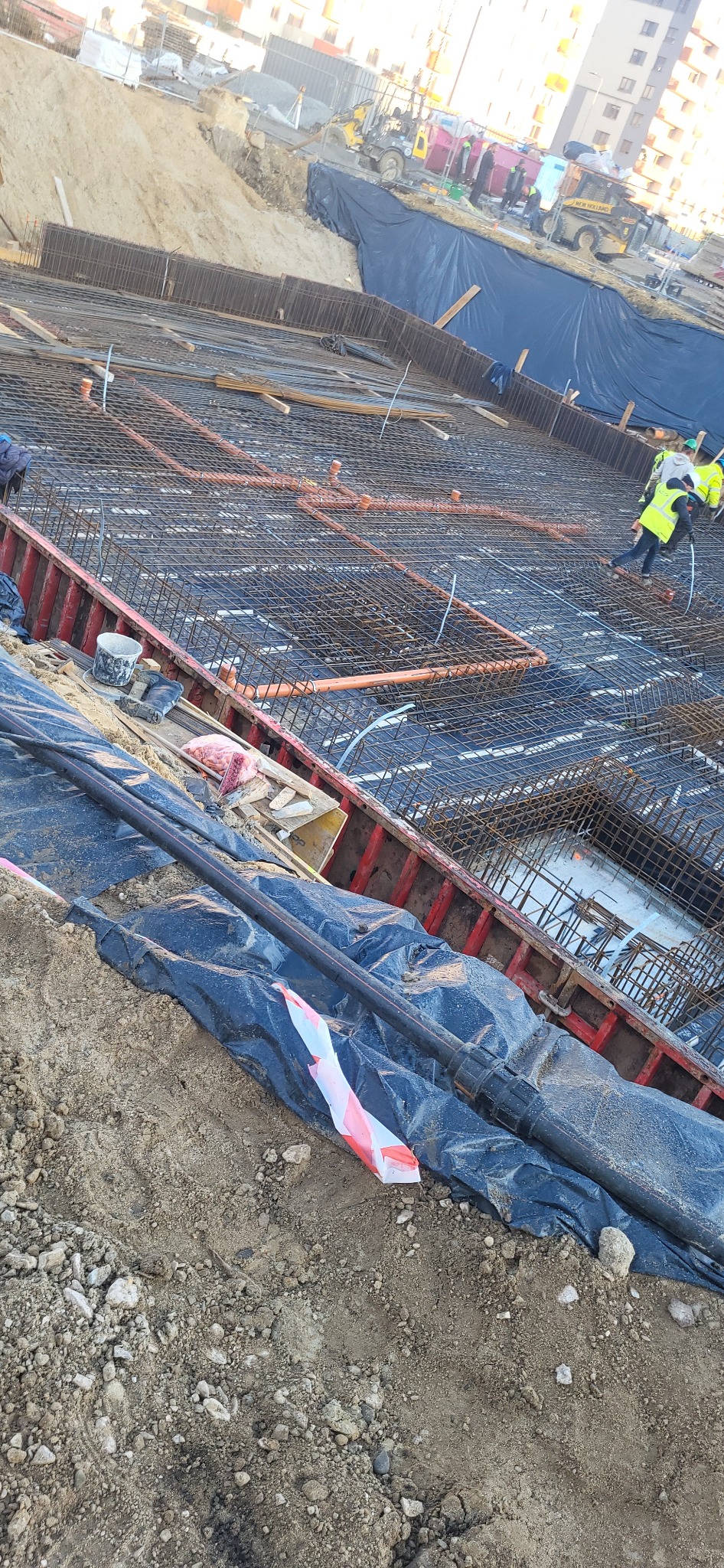 Budowa fundamentów budynku z widocznym zbrojeniem, rurami instalacyjnymi i pracownikami w kamizelkach odblaskowych na placu budowy.