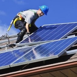 M-tech Mariusz Bartosz - Montaż paneli fotowoltaicznych na dachu domu przez instalatora w niebieskim kasku, zabezpieczonego uprzężą, na tle błękitnego nieba.