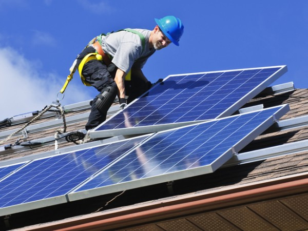 Montaż paneli fotowoltaicznych na dachu domu przez instalatora w niebieskim kasku, zabezpieczonego uprzężą, na tle błękitnego nieba.