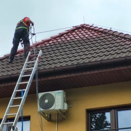 Totall Clean - Pracownik w uprzęży na drabinie, częściowo pomalowany dach z brązowej dachówki, żółta elewacja z klimatyzatorem i oknami.