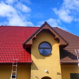 Totall Clean - Dwukolorowy dach z dachówki ceramicznej, częściowo pomalowany na czerwono, z aluminiowymi drabinami opartymi o ścianę budynku z okrągłym oknem, na tle błękitnego nieba z chmurami.