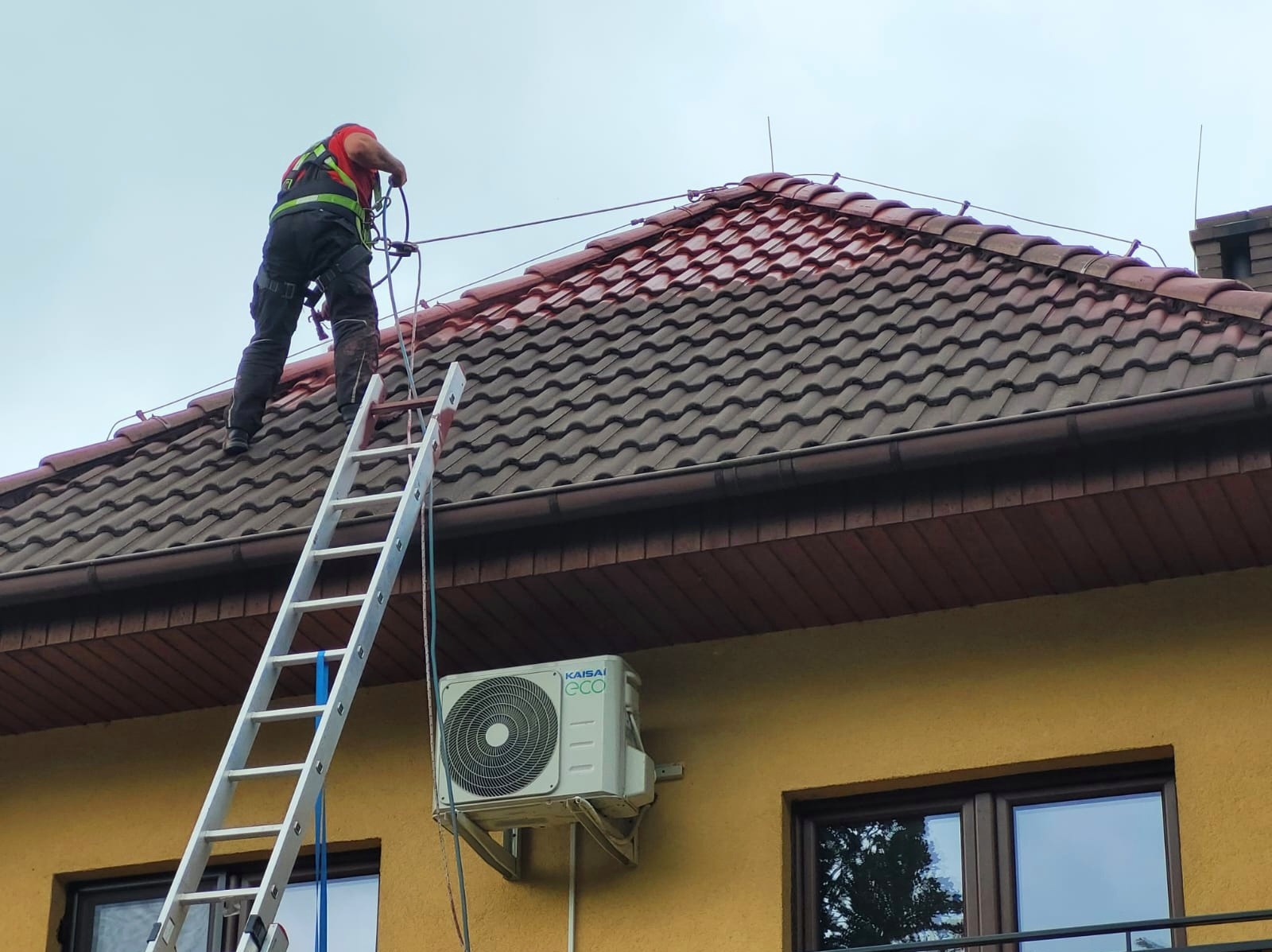 Pracownik w uprzęży na drabinie, częściowo pomalowany dach z brązowej dachówki, żółta elewacja z klimatyzatorem i oknami.