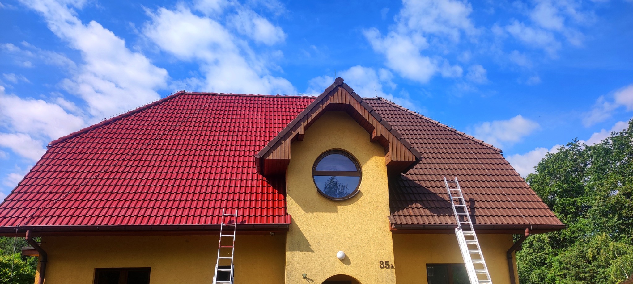 Dwukolorowy dach z dachówki ceramicznej, częściowo pomalowany na czerwono, z aluminiowymi drabinami opartymi o ścianę budynku z okrągłym oknem, na tle błękitnego nieba z chmurami.