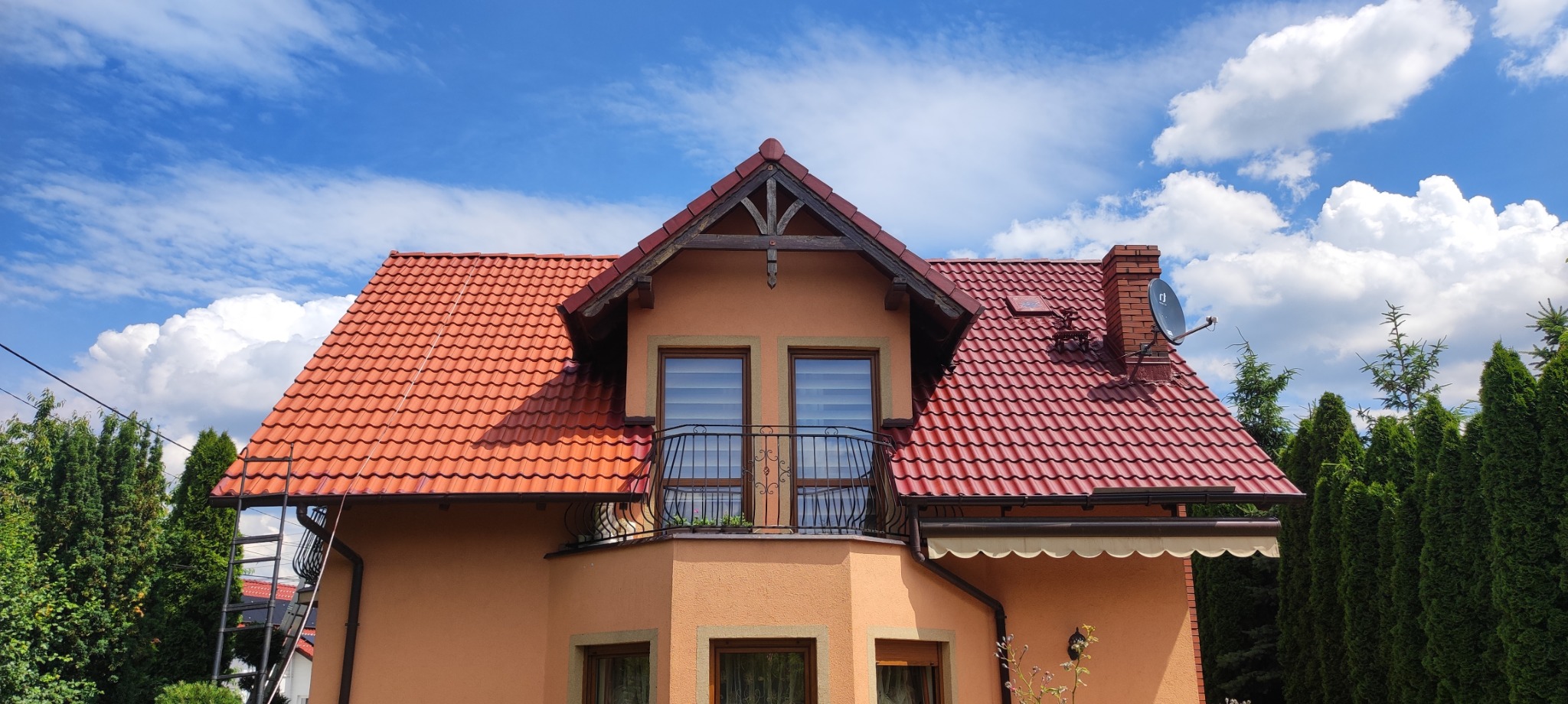 Dom jednorodzinny z nową, czerwoną dachówką, balkonem z ozdobną balustradą i elewacją w odcieniach beżu, na tle błękitnego nieba z białymi chmurami