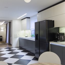 Dom Pracy Twórczej Łukasz Engel - Nowoczesna kuchnia z czarno-białą podłogą w szachownicę, szarymi szafkami i białymi blatami z marmurowym wzorem, czarną lodówką i ekspresem do kawy, widok na salon z fortepianem w tle.