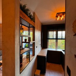 Dom Pracy Twórczej Łukasz Engel - Ciemna kuchnia z oknem i drewnianą półką z roślinami, widok z korytarza, ciepłe oświetlenie sufitowe.