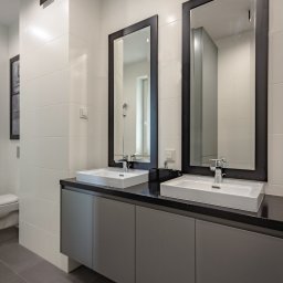 Dom Pracy Twórczej Łukasz Engel - Nowoczesna łazienka z dwoma umywalkami nablatowymi, czarnym blatem i ramami luster, szara szafka pod umywalkami, białe płytki na ścianach i szara podłoga, w tle widoczna toaleta i obraz na ścianie.