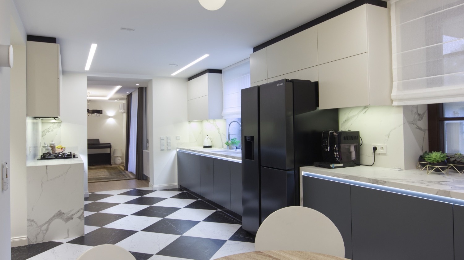 Nowoczesna kuchnia z czarno-białą podłogą w szachownicę, szarymi szafkami i białymi blatami z marmurowym wzorem, czarną lodówką i ekspresem do kawy, widok na salon z fortepianem w tle.
