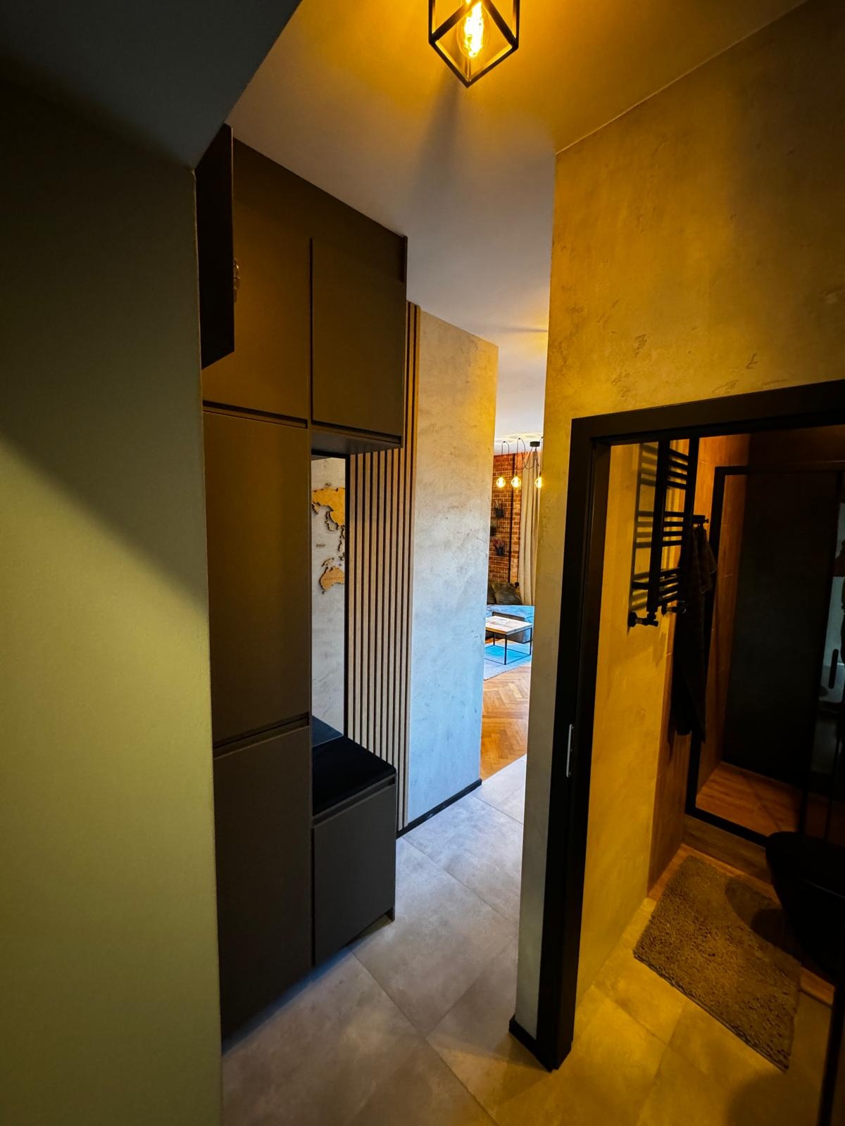 Wnętrze mieszkania: widok z korytarza na salon i łazienkę, z zabudowaną szafą w przedpokoju, dekoracyjną ścianą z drewnianymi lamelami i mapą świata.