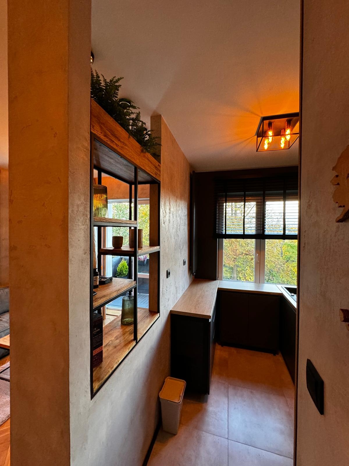 Ciemna kuchnia z oknem i drewnianą półką z roślinami, widok z korytarza, ciepłe oświetlenie sufitowe.