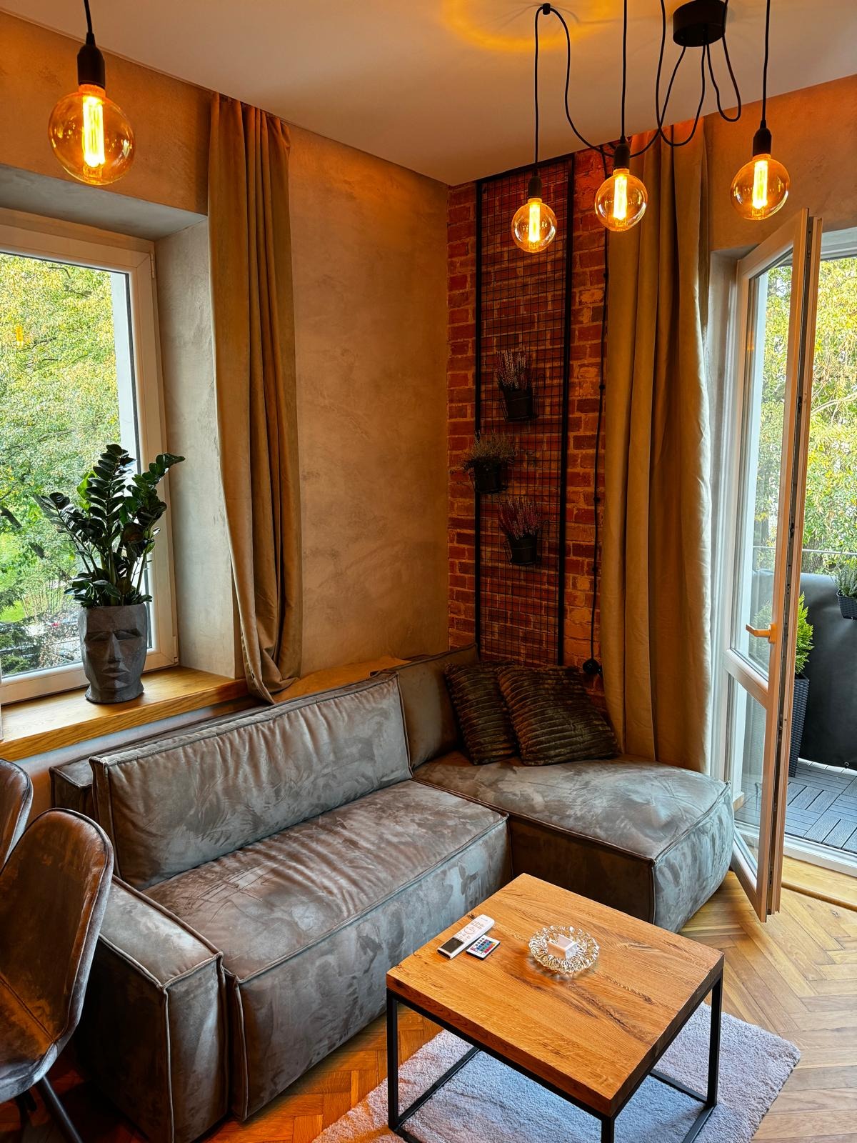 Salon z szarą, modułową sofą narożną, drewnianym stolikiem kawowym z popielniczką, rośliną w doniczce z motywem twarzy na parapecie i lampami wiszącymi z żarówkami Edisona.