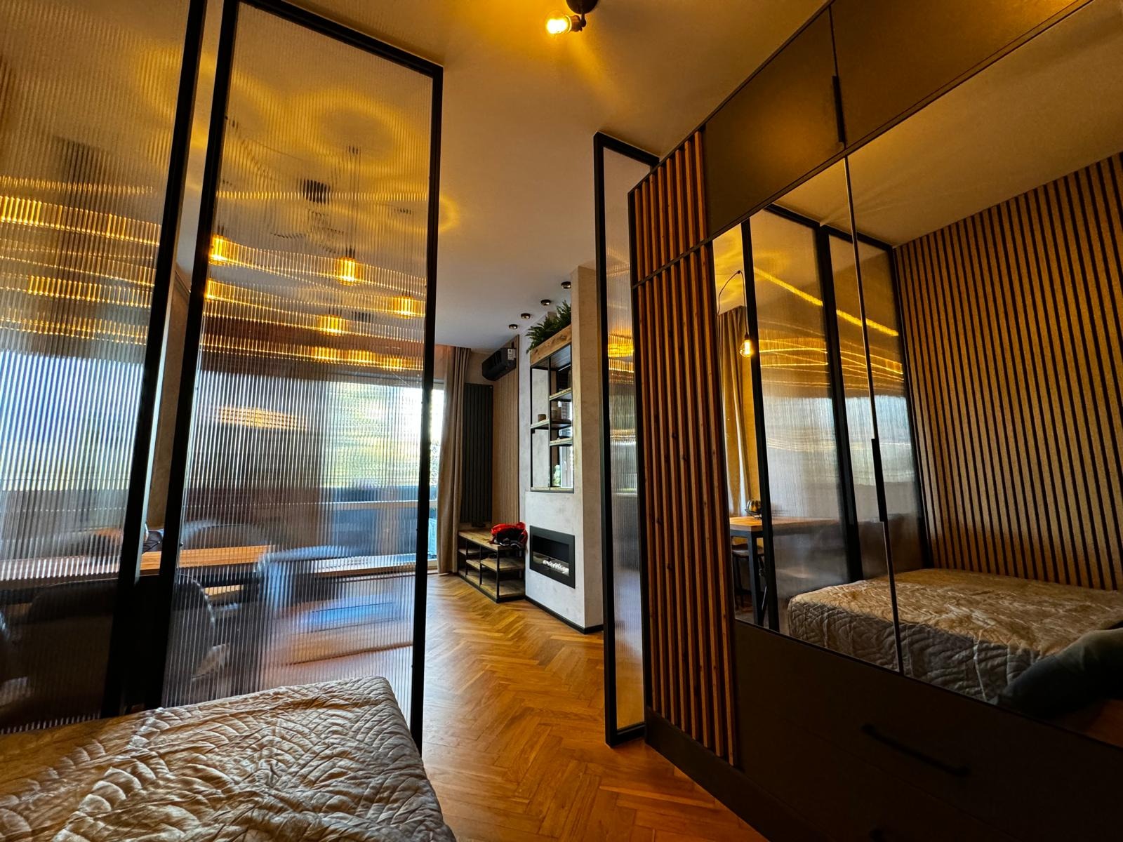 Nowoczesne wnętrze mieszkania z podziałem przestrzeni za pomocą szklanych ścianek działowych i drewnianych paneli, parkiet w jodełkę, widoczny kominek elektryczny i elementy dekoracyjne.