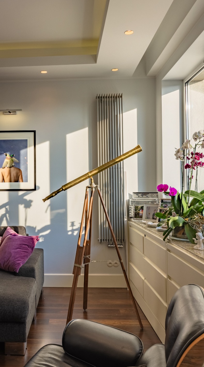 Salon z drewnianą podłogą, szarą sofą z fioletową poduszką, teleskopem na statywie, grzejnikiem żeberkowym i białymi szafkami pod oknem z kwiatami.