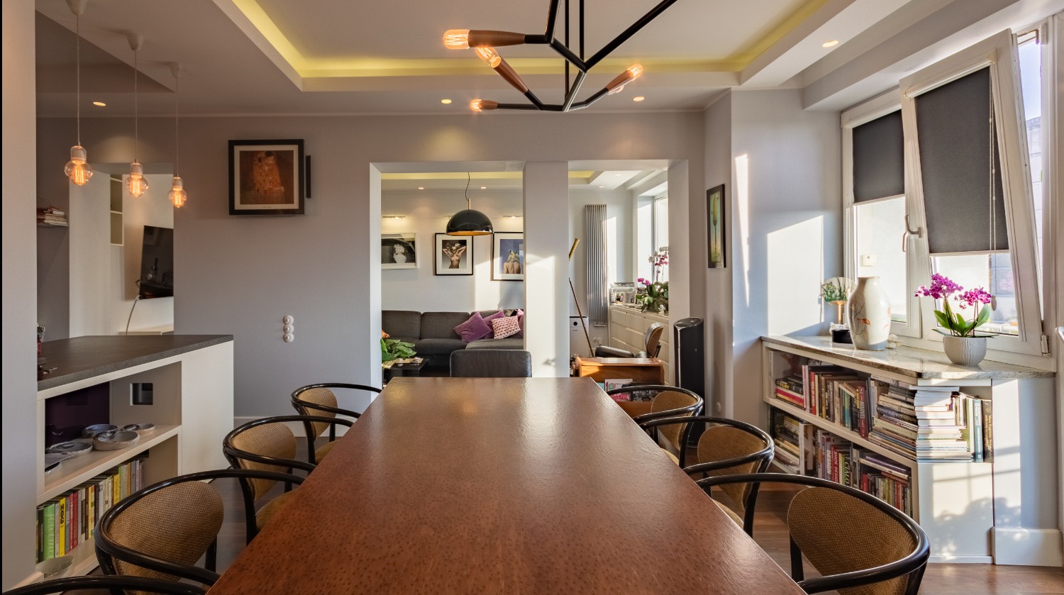 Przestronne wnętrze mieszkania z dużym drewnianym stołem, krzesłami z wikliny, widokiem na salon przez otwarte przejście i oświetleniem sufitowym w stylu industrialnym.