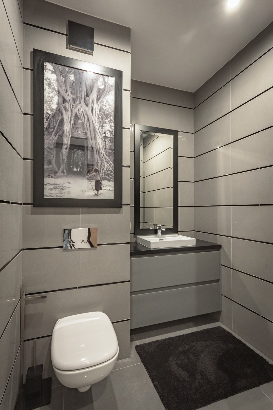 Nowoczesna łazienka w odcieniach szarości z poziomymi płytkami, wiszącą toaletą, szafką z umywalką i czarnym dywanikiem; na ścianie wisi oprawiona w ramę fotografia Angkor Wat.