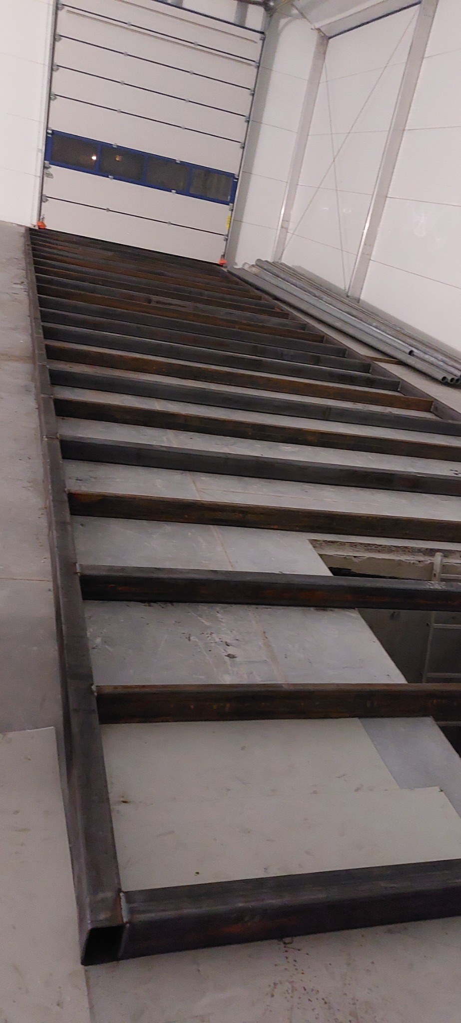 Konstrukcja stalowa podestu z profili zamkniętych w hali produkcyjnej, widok z dołu na bramę segmentową w tle.