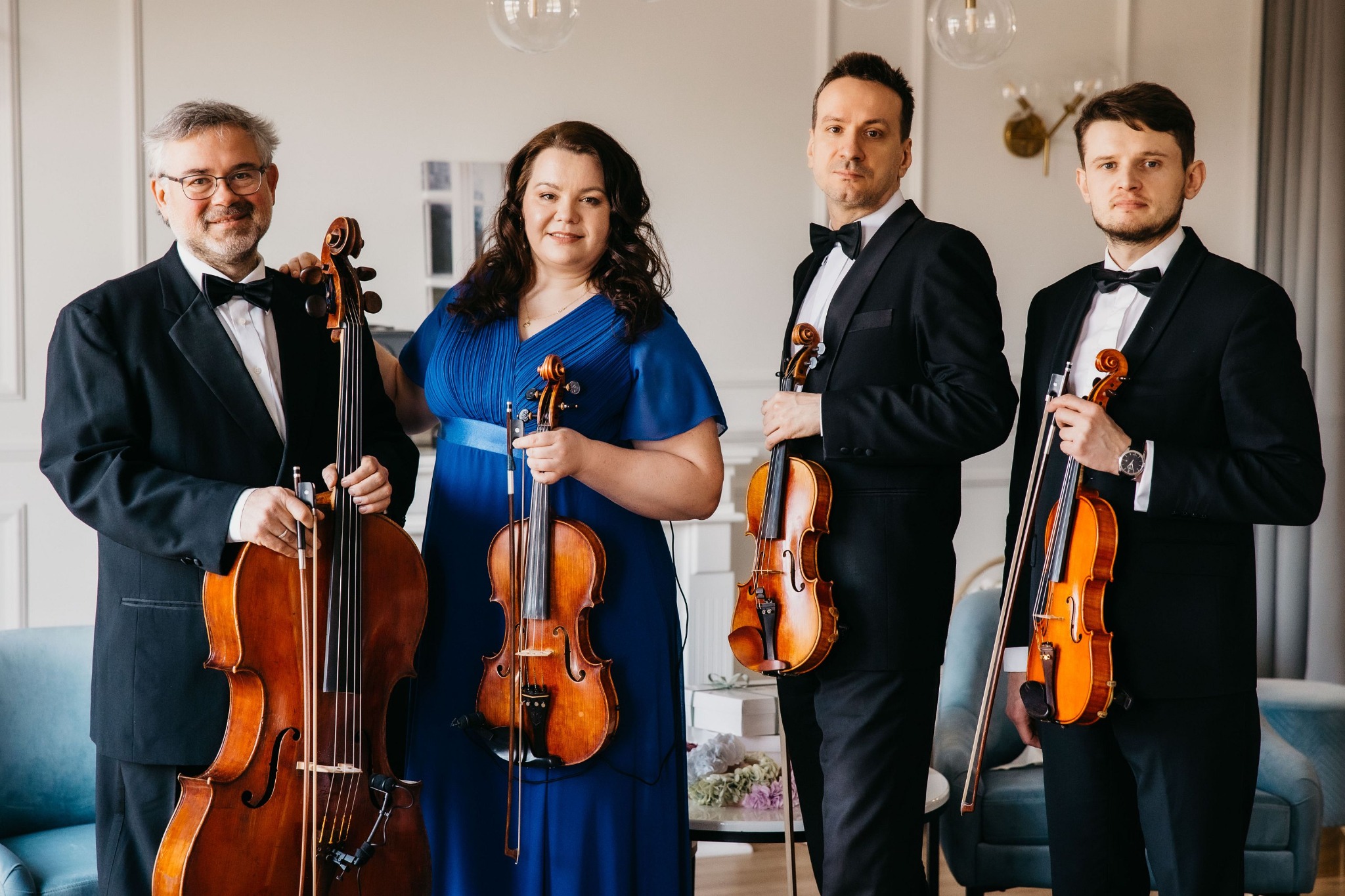 Kwartet smyczkowy w formalnych strojach, trzymający skrzypce i wiolonczelę, pozujący w jasnym wnętrzu z dekoracyjnymi lampami.