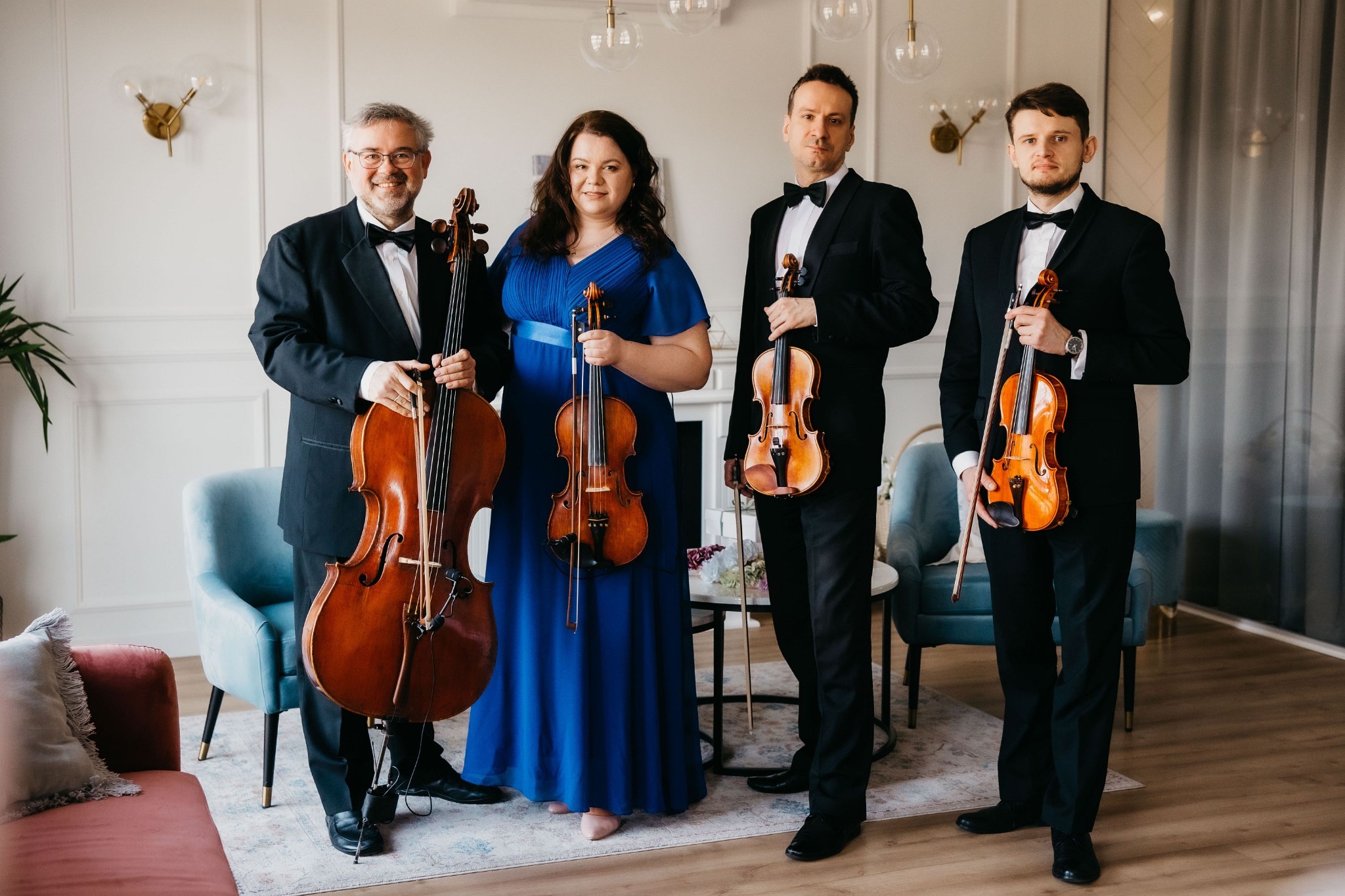 Kwartet smyczkowy w formalnych strojach pozuje z instrumentami: wiolonczelą i skrzypcami, w jasnym wnętrzu z dekoracyjnymi lampami.