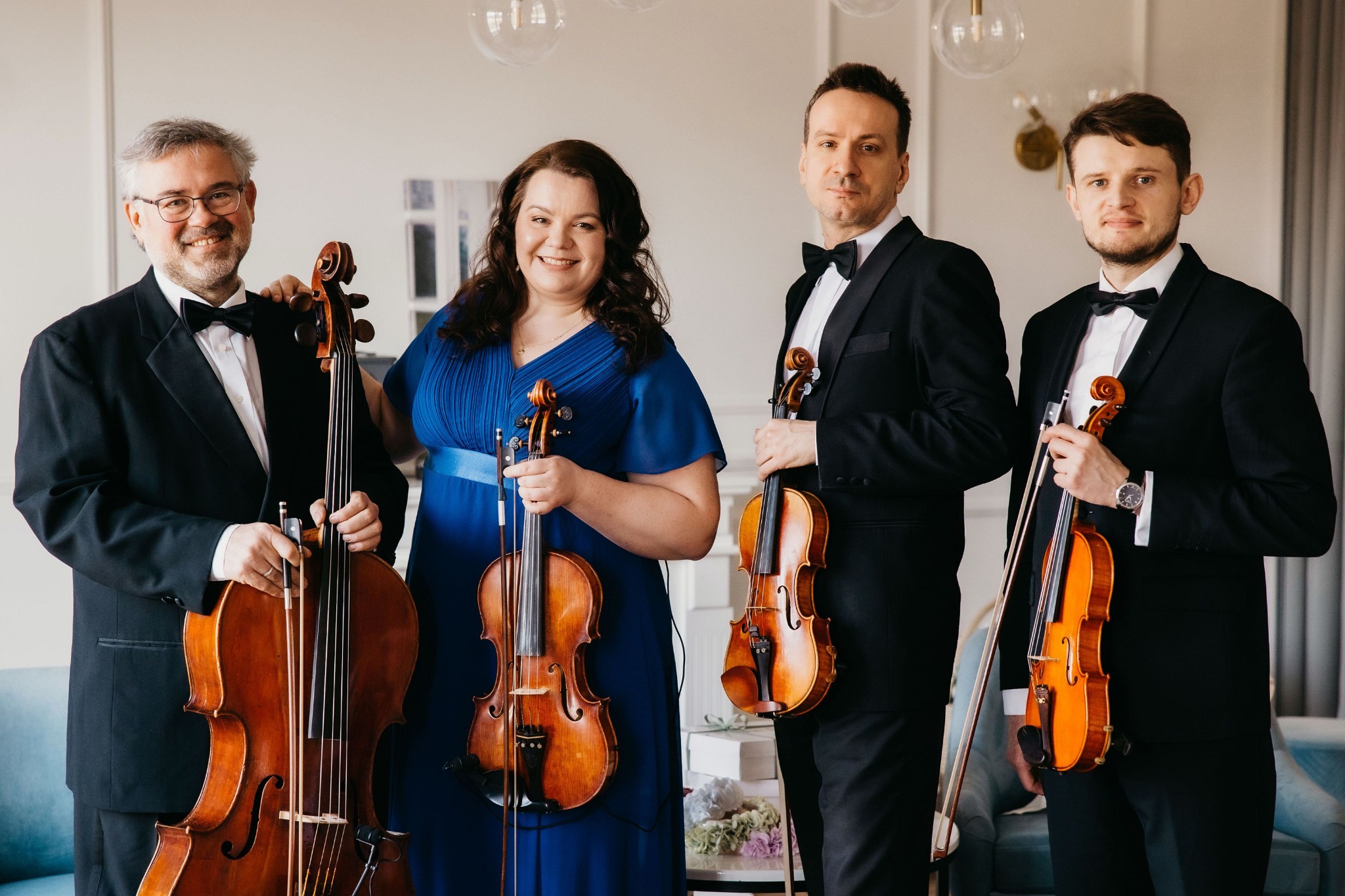 Kwartet smyczkowy: uśmiechnięci muzycy w smokingach i granatowej sukni prezentują skrzypce i wiolonczelę w jasnym wnętrzu.