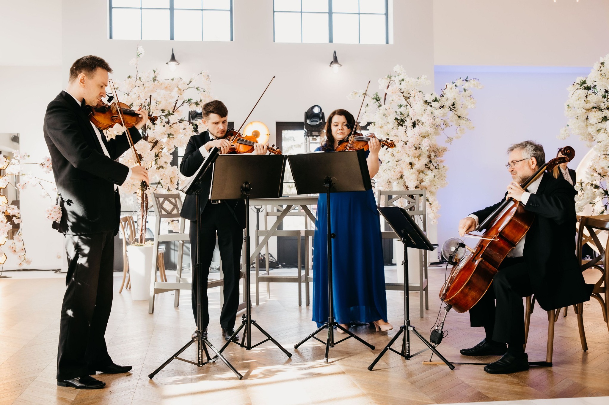 Kwartet smyczkowy: Dwóch skrzypków, altowiolistka w długiej, błękitnej sukni i wiolonczelista w eleganckim stroju grający na tle dekoracji z białych kwiatów.