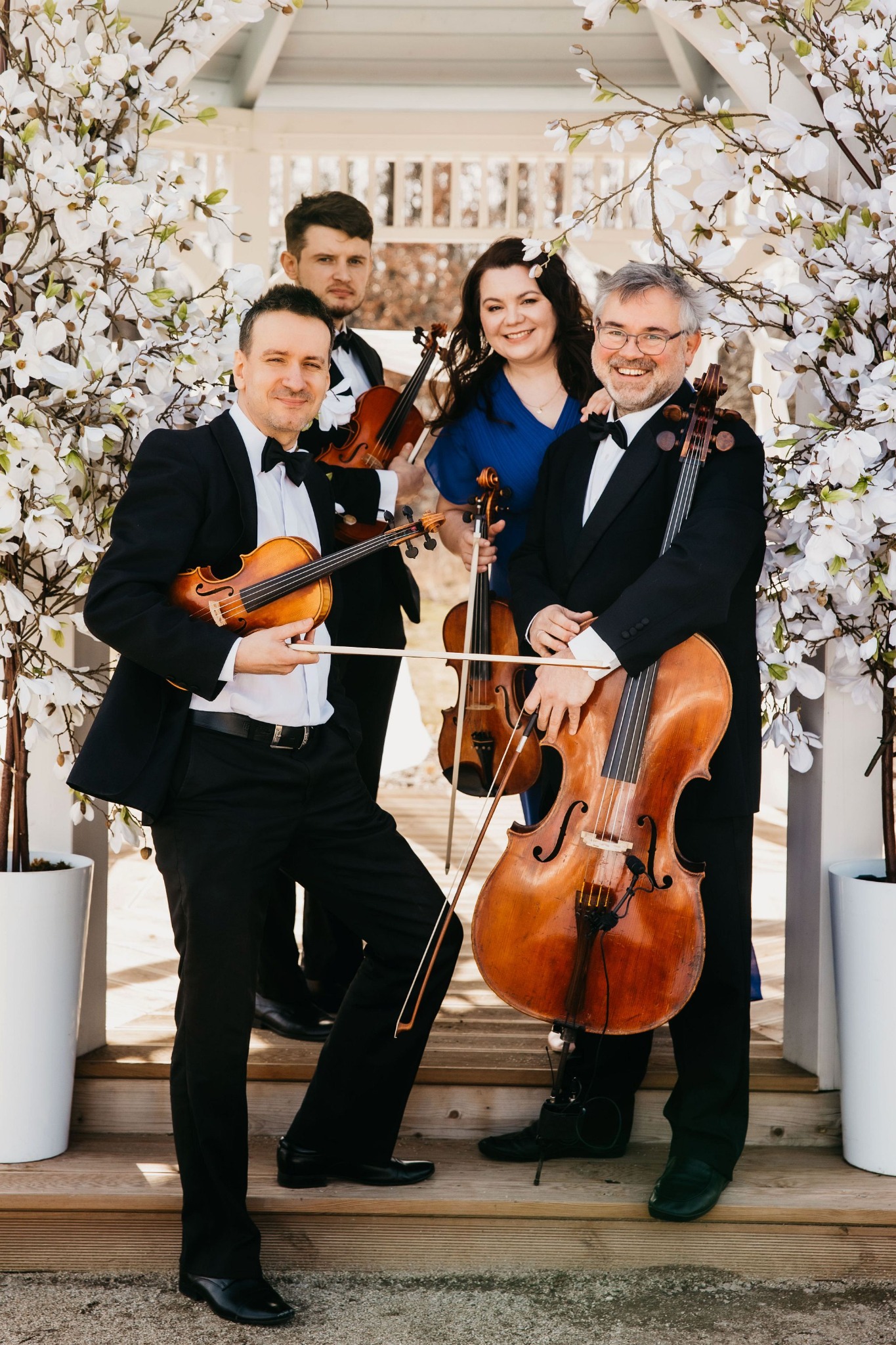 Kwartet smyczkowy w formalnych strojach pozuje z instrumentami przed białym altaną udekorowaną kwitnącymi gałązkami.