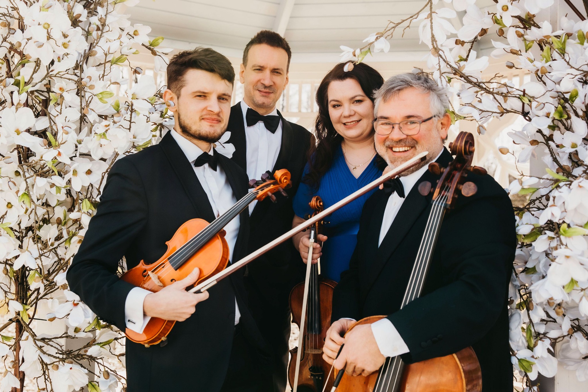 Kwartet smyczkowy w smokingach pozuje z instrumentami na tle białych kwiatów.