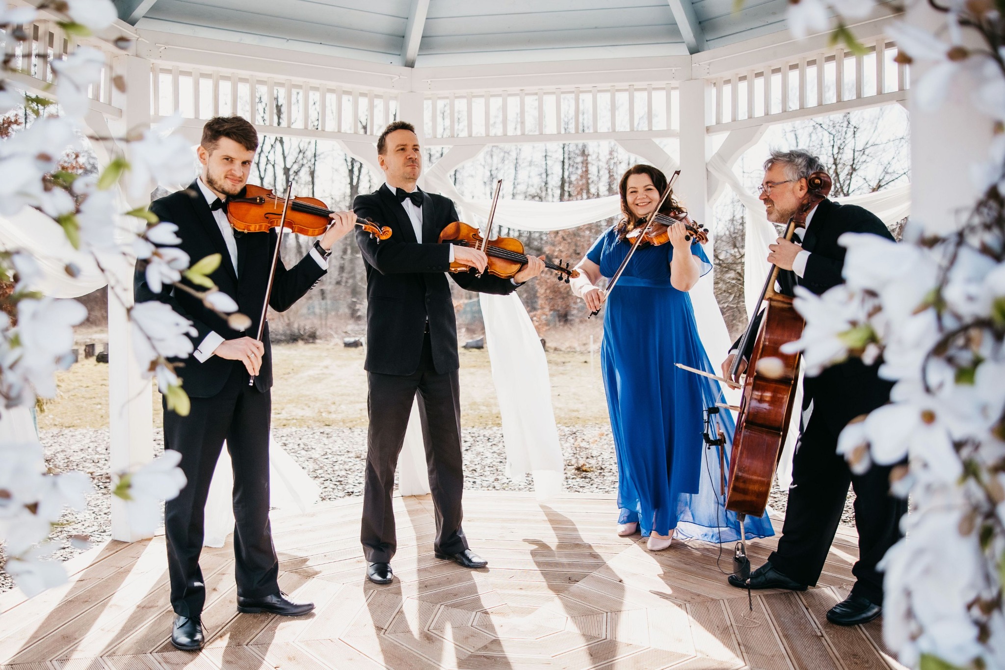 Kwartet smyczkowy w strojach wieczorowych gra w białej altanie udekorowanej kwitnącymi gałązkami magnolii.
