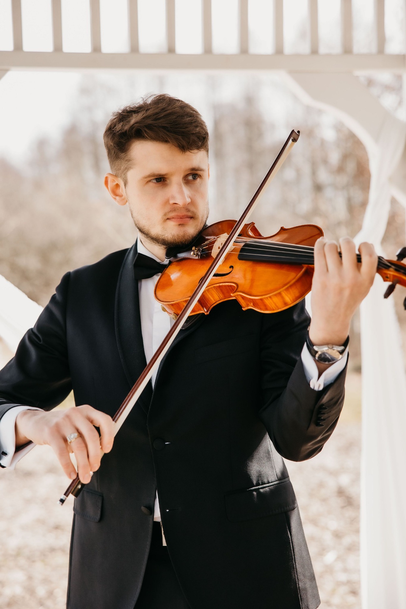 Elegancko ubrany skrzypek w muszce, grający na skrzypcach pod białą altaną, z widocznym pierścieniem na palcu i zegarkiem na ręce.